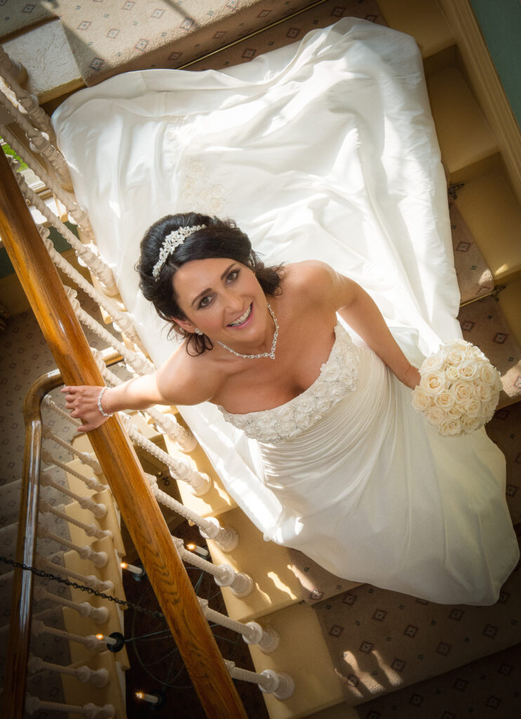 High-angle shot of a bride in a strapless floral-accented wedding gown looking up from the historic staircase at Weetwood Hall.