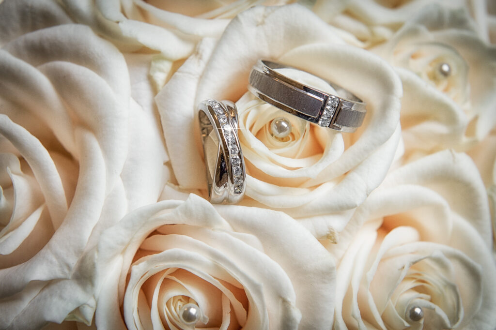 Close-up of two silver wedding bands resting inside the petals of white roses, with a focus on the diamond detailing.