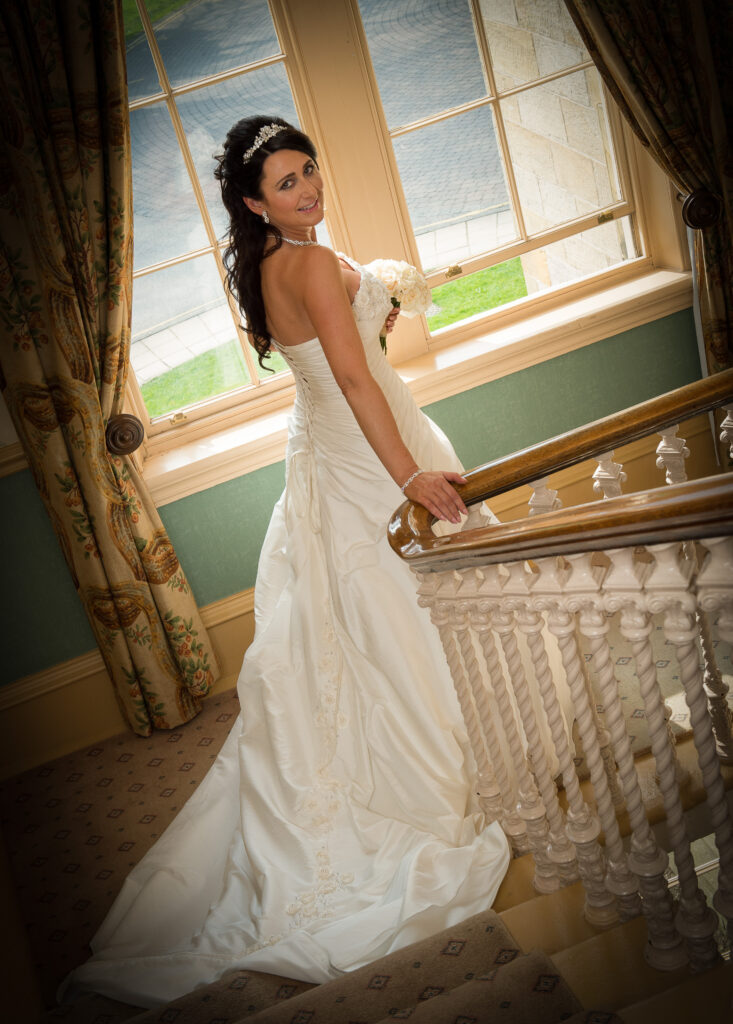 Bride standing by a large manor window at Weetwood Hall, showing the back detail of her gown and holding a bouquet.