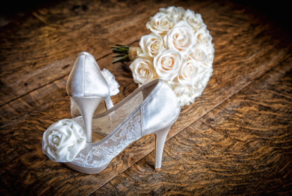 A pair of white lace and satin bridal heels next to a white rose bouquet on a rustic wooden table at Weetwood Hall.