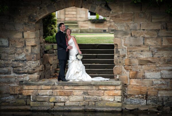 A bride and groom laughing together, framed by a rustic stone archway at a historic Leeds wedding estate. The reflection of the stone is visible in the water below.