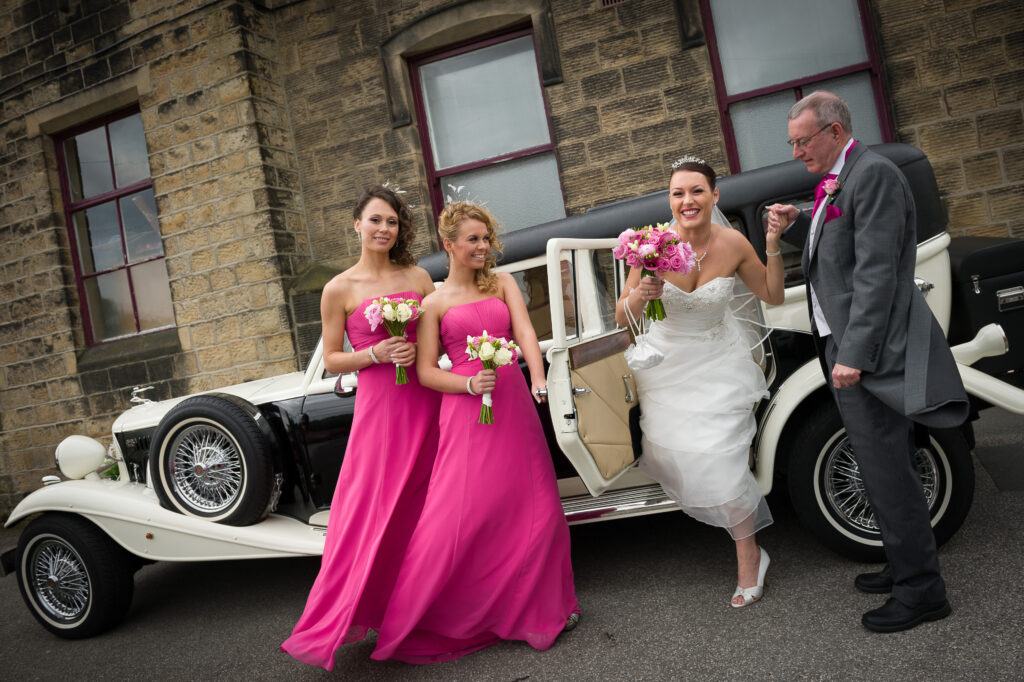 A bride in a tiered white gown stepping out of a vintage white wedding car with bridesmaids in hot pink dresses at a West Yorkshire estate.