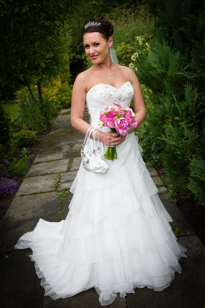 A bride wearing a tiered white wedding dress and tiara holding a pink rose and calla lily bouquet in a lush garden setting.