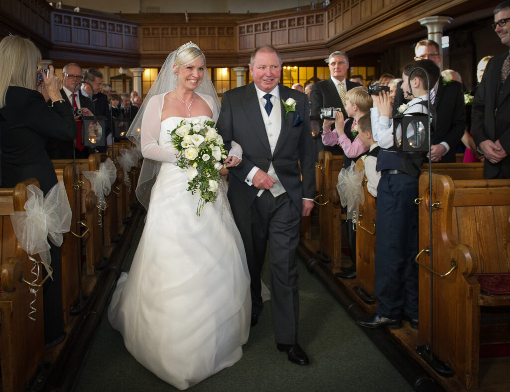 The bride and her father walking down the aisle of Christ Church Harrogate, lined with lanterns and white bows.
