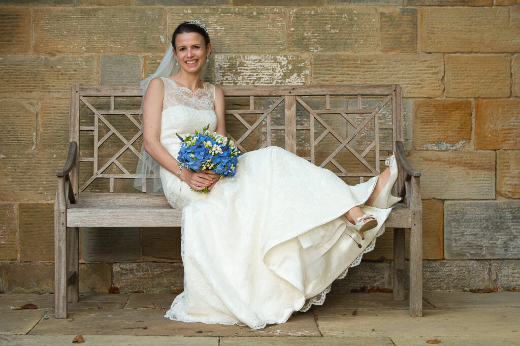 Bride in lace wedding dress sitting on a wooden bench with blue bouquet at Rudding Park Howard Barnett Photography