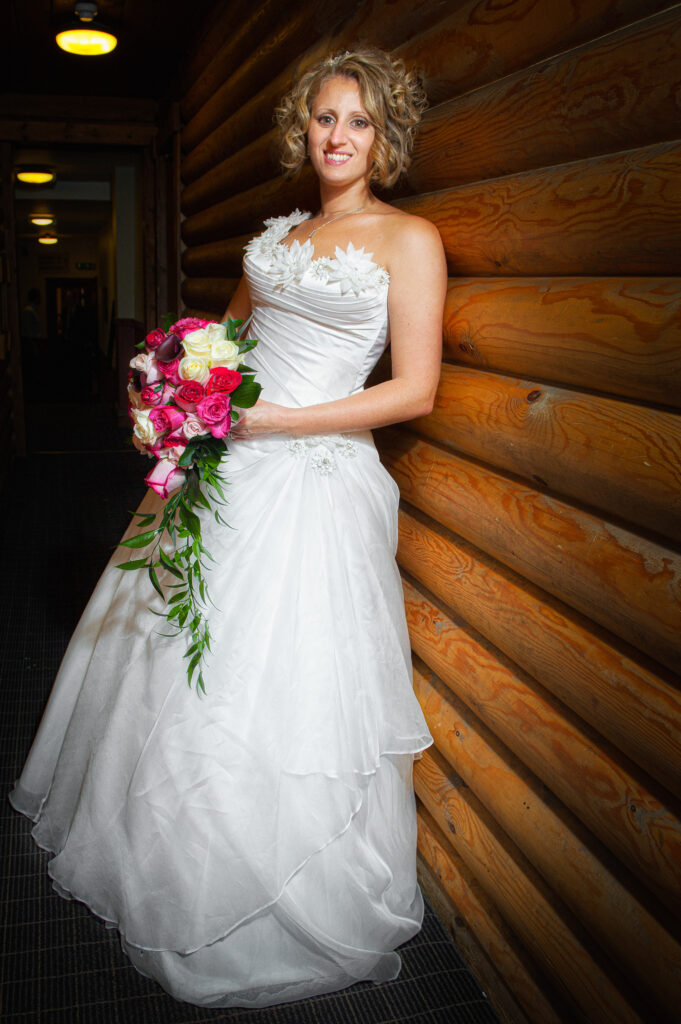 A bride in a white wedding dress holding a pink and cream rose bouquet leaning against a rustic log wall at Chevin Lodge.
