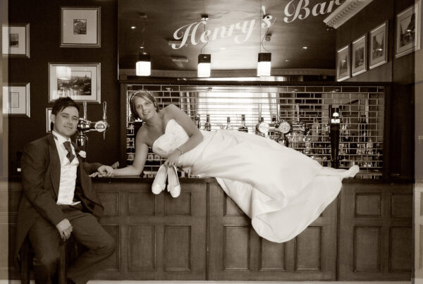 Sepia-toned photo of a bride laying across the counter at Henry’s Bar in The Crown Hotel, Harrogate, holding her shoes, with the groom sitting next to her.