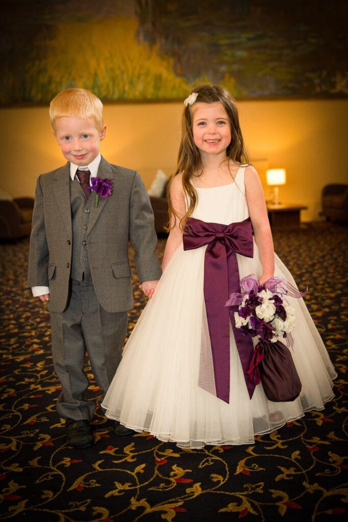 A young flower girl in a white dress with a purple sash and a page boy in a grey suit holding hands at a wedding in Leeds.