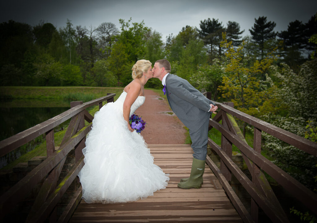 A bride and groom leaning in for a kiss on a rustic wooden bridge with a lush green Yorkshire background.
