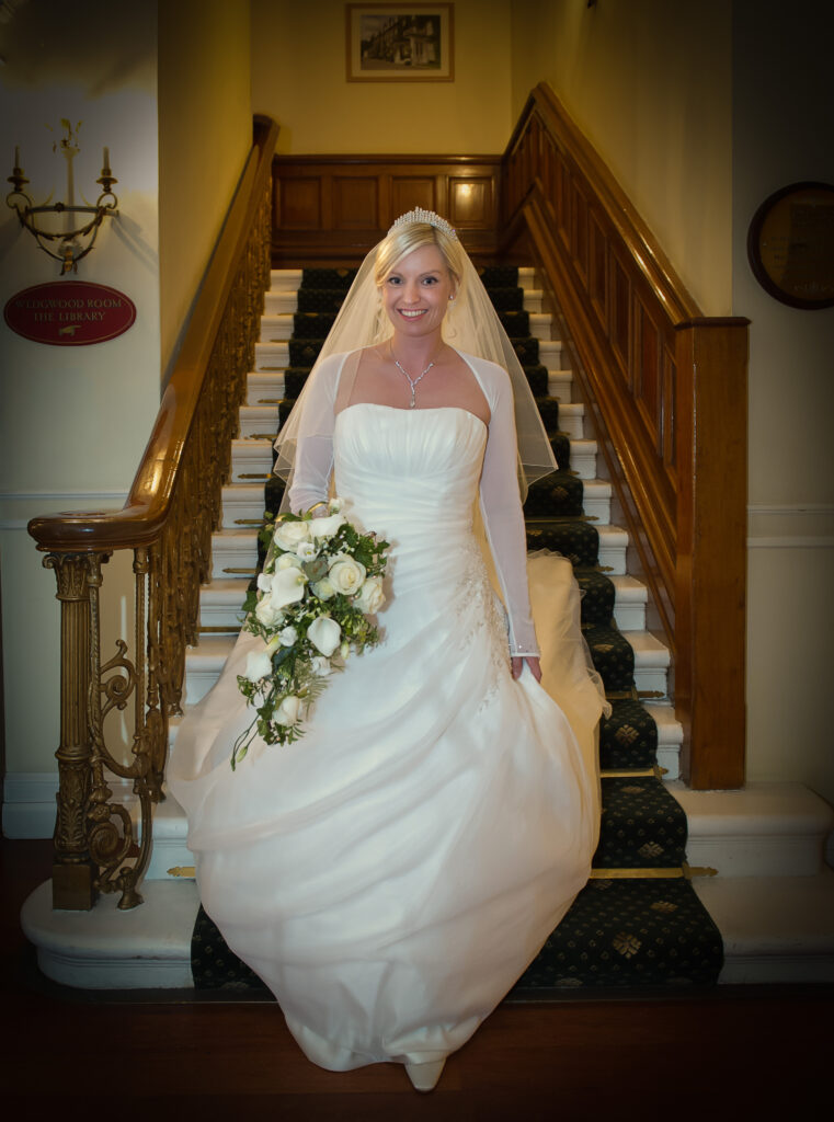 A smiling bride in a strapless white wedding gown standing on the grand wooden staircase at The Old Swan Hotel Harrogate.