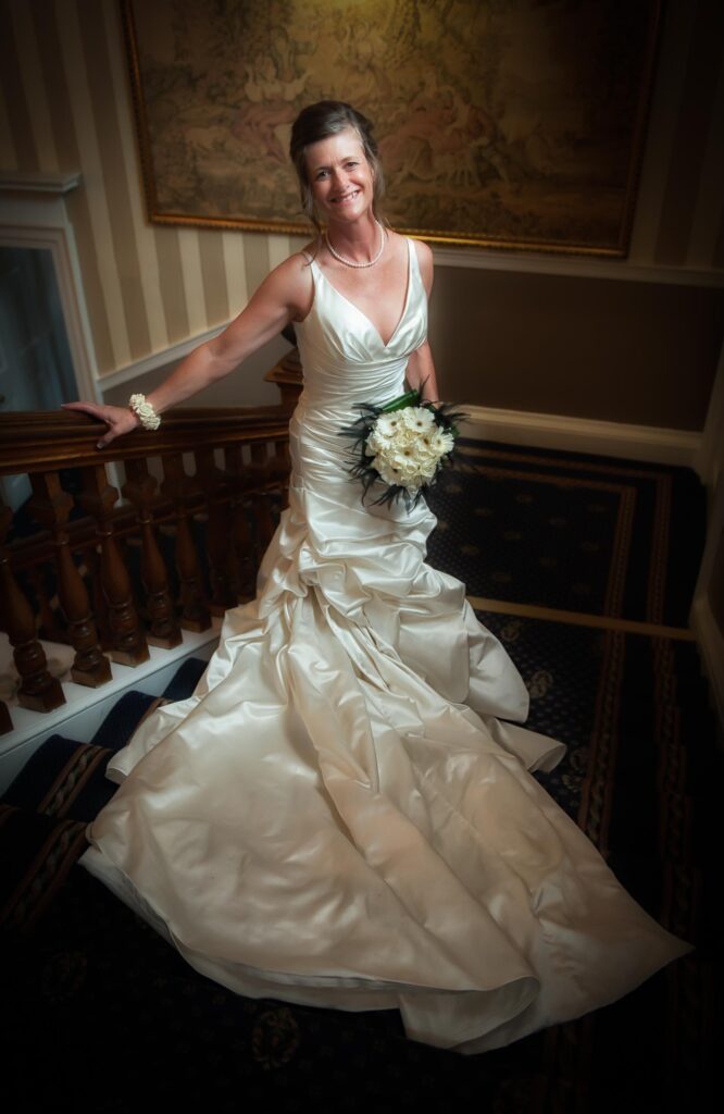 A stunning bride in a silk ruched wedding dress posing on a grand wooden staircase in a Leeds wedding venue. She is holding a white gerbera daisy bouquet with black feather accents.
