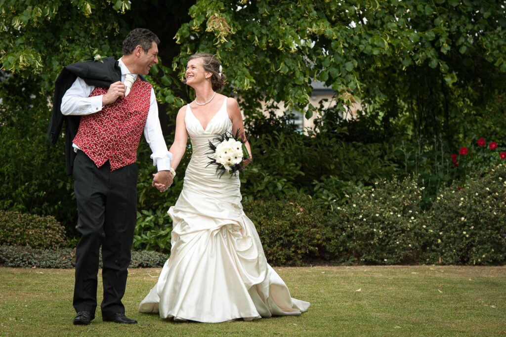 A happy bride and groom walking hand-in-hand through a lush green garden in West Yorkshire. The groom is wearing a red patterned waistcoat and carrying his suit jacket over his shoulder.