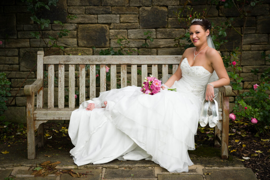 bride sitting comfortably on a weathered wooden bench against a stone wall, holding her shoes and a pink bouquet.