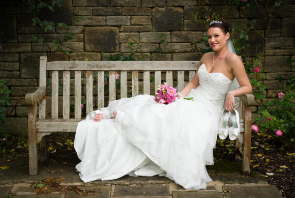 bride sitting comfortably on a weathered wooden bench against a stone wall, holding her shoes and a pink bouquet.