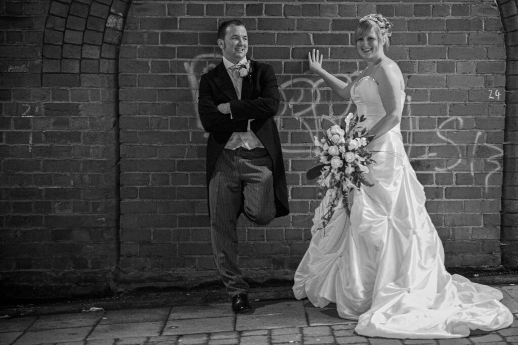 A relaxed and creative portrait of a bride and groom leaning against a rustic brick wall in an urban Leeds setting, captured by Howard Barnett Photography.
