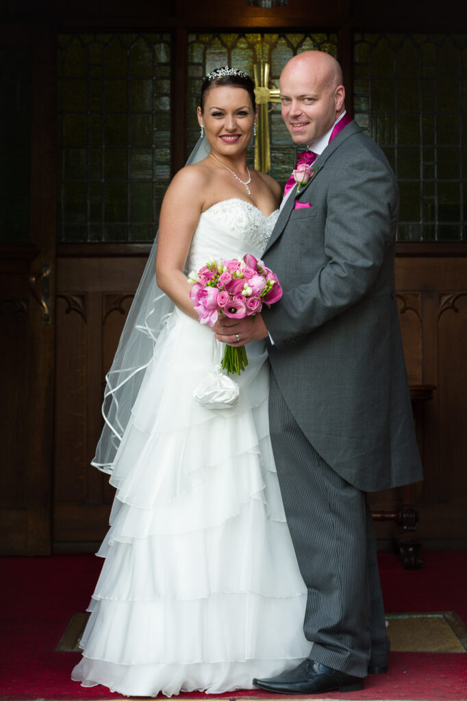 A bride and groom posing together in front of a dark wood doorway with a gold cross at a West Yorkshire wedding venue.