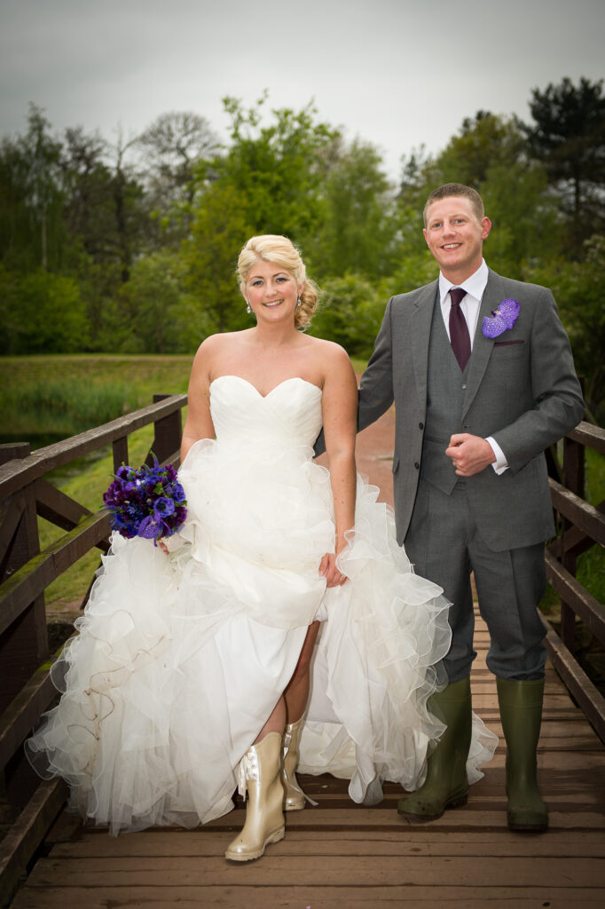 A bride lifts her gown to show off cream wedding wellies while standing on a wooden bridge with her groom in Yorkshire.