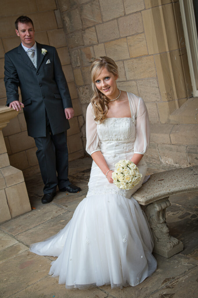 Elegant bride in a lace wedding dress sitting on a historic stone bench at Weetwood Hall Estate in Leeds.