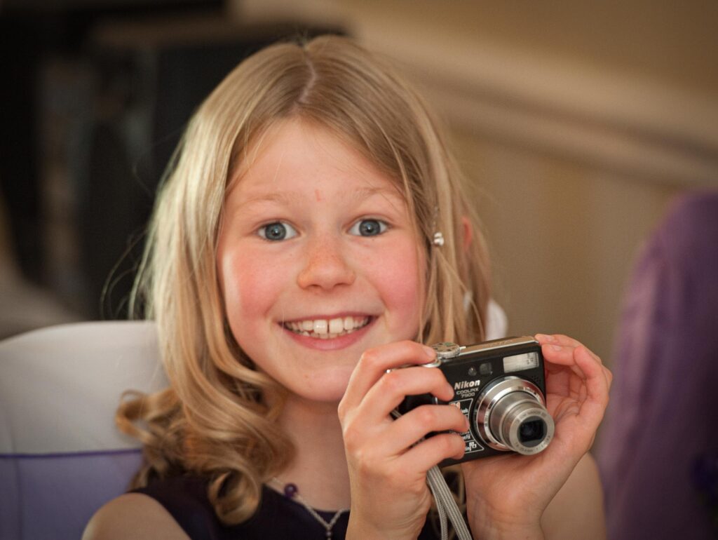 A young guest smiling and holding a digital camera at a Leeds wedding venue, photographed by Howard Barnett.