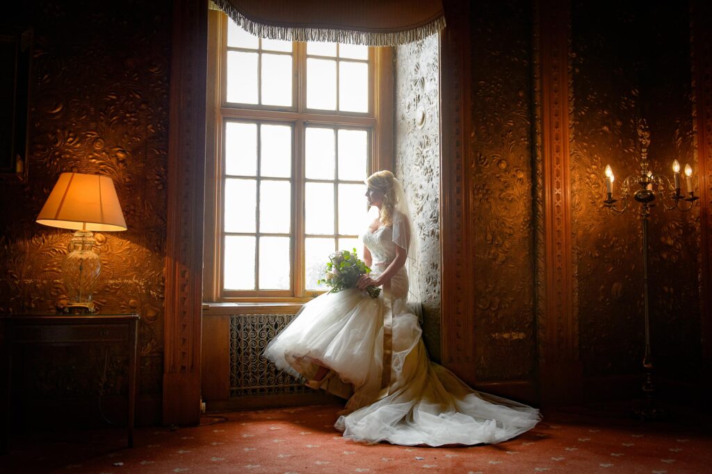 A close-up side profile of a bride in a veiled dress with a small bouquet, looking out a large, multi-paned window. Soft natural light illuminates her, by a Leeds wedding photographer.