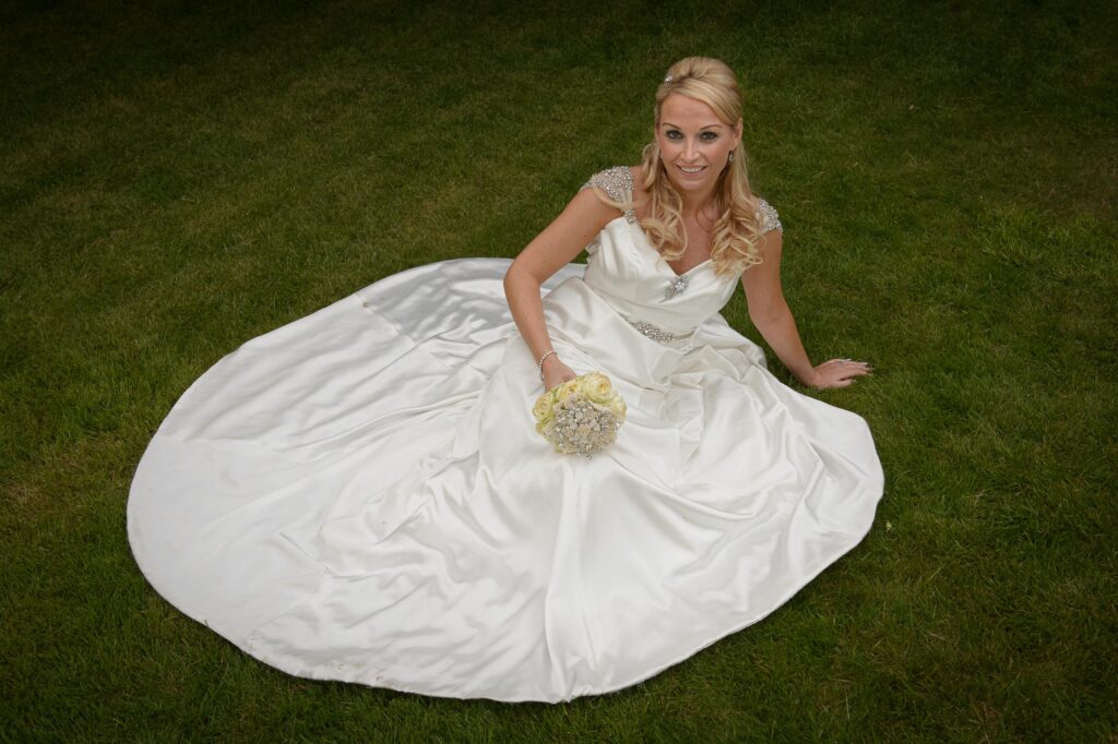 Bride on Grass: "A bride in a white satin wedding dress sitting on the lawn at The Old Swan in Harrogate, holding a rose bouquet."