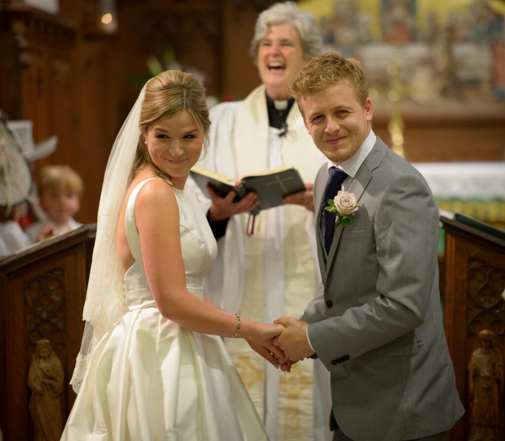 Bride and groom holding hands during a church wedding ceremony with a smiling officiant, Leeds wedding photographer.
