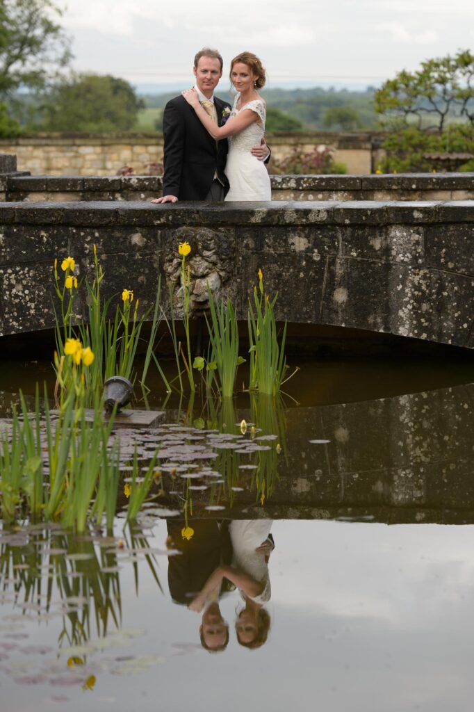 A creative wedding photo by Howard Barnett showing a bride and groom leaning over a stone bridge, with their reflection visible in the pond water below.