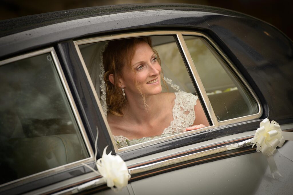 Close-up bridal portrait by Howard Barnett showing a bride with a lace veil looking out of a vintage car window decorated with white bows.