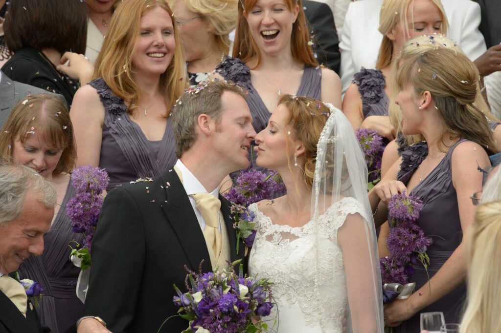 A bride and groom kissing while surrounded by bridesmaids in purple dresses holding allium bouquets as confetti falls around them.