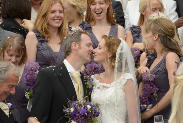 A bride and groom kissing while surrounded by bridesmaids in purple dresses holding allium bouquets as confetti falls around them.