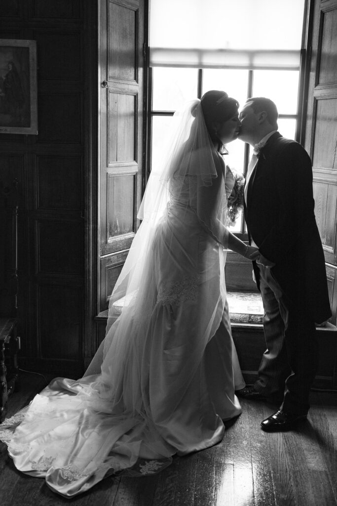 Black and white wedding photography of a bride and groom sharing a kiss by a window, captured by Howard Barnett.