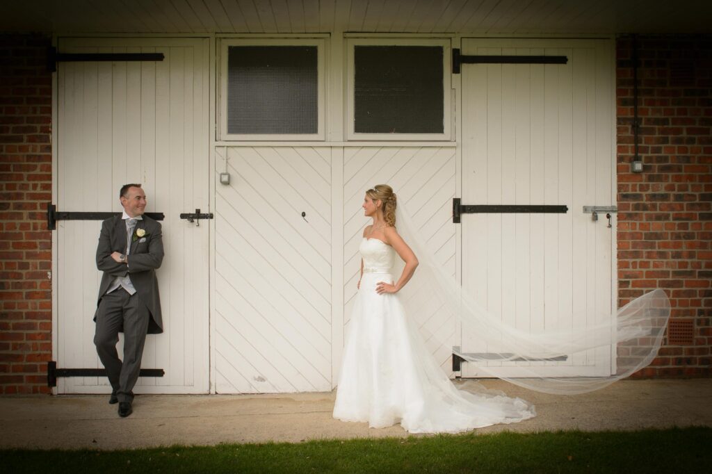 Creative wedding portrait of a bride with a long flying veil and groom leaning against white barn doors in West Yorkshire.