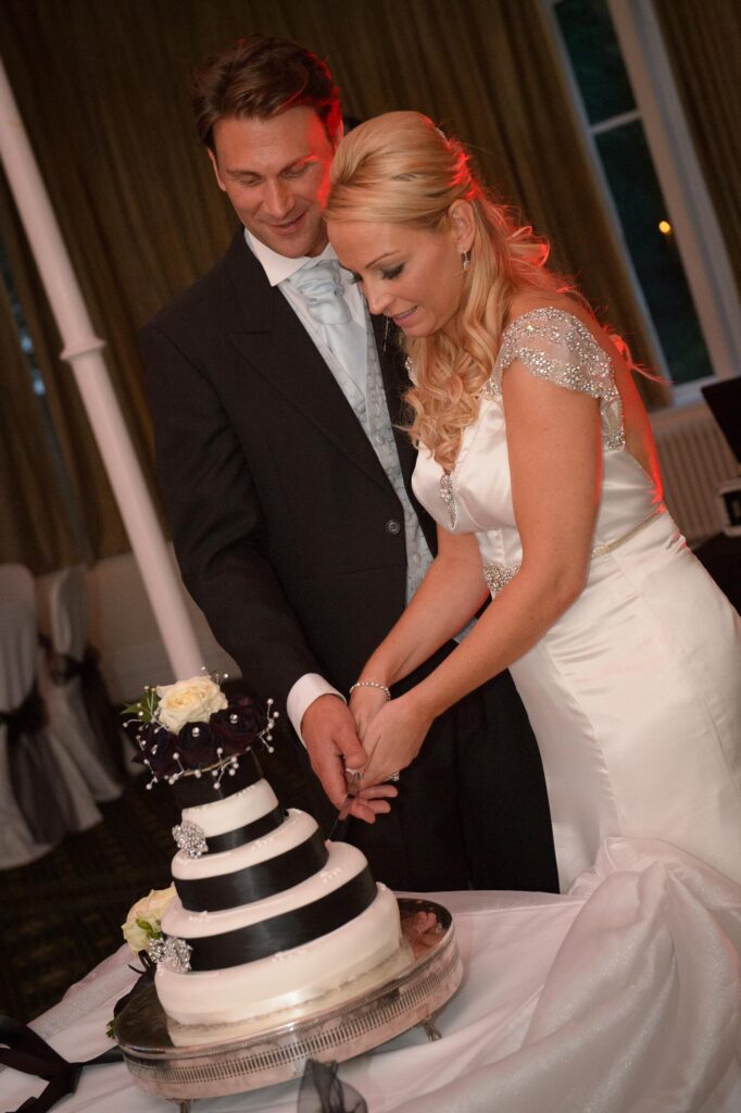 Cake Cutting: "A bride and groom smiling while cutting their black and white tiered wedding cake at a Leeds reception, captured by Howard Barnett Photography."