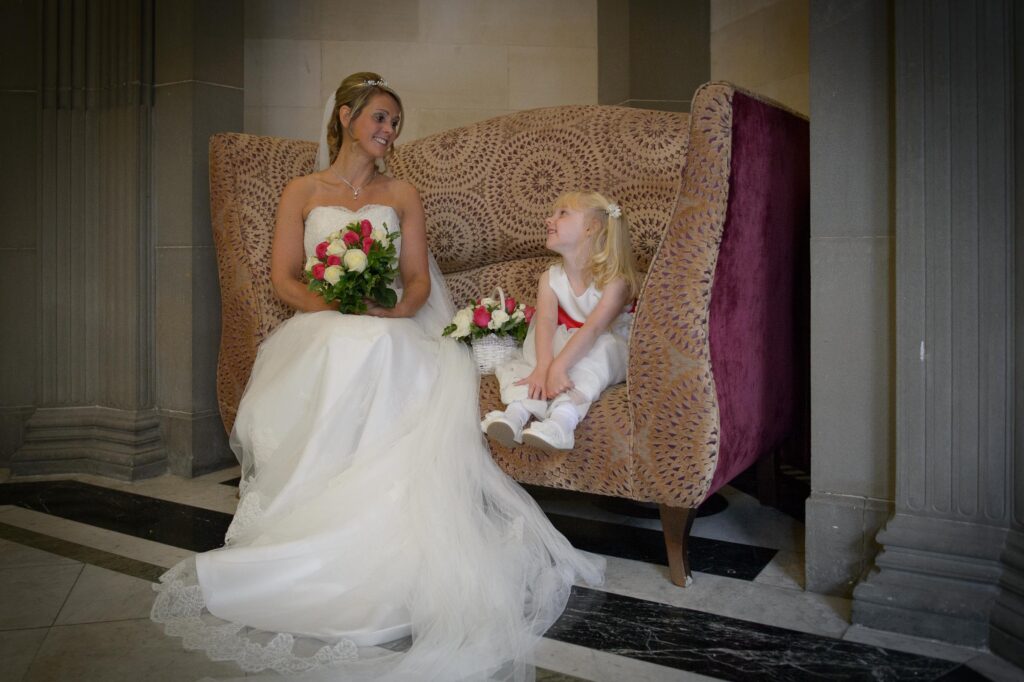 Candid moment of a bride sitting on a vintage sofa talking to a young flower girl, captured by Howard Barnett Photography, Leeds.