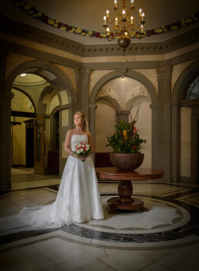 Atmospheric wide shot of a bride standing in a grand architectural hall with a chandelier and floral centerpiece in West Yorkshire.