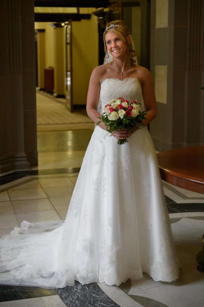 Full-length bridal portrait of a bride holding a rose bouquet in a grand Leeds venue hallway, photographed by Howard Barnett.