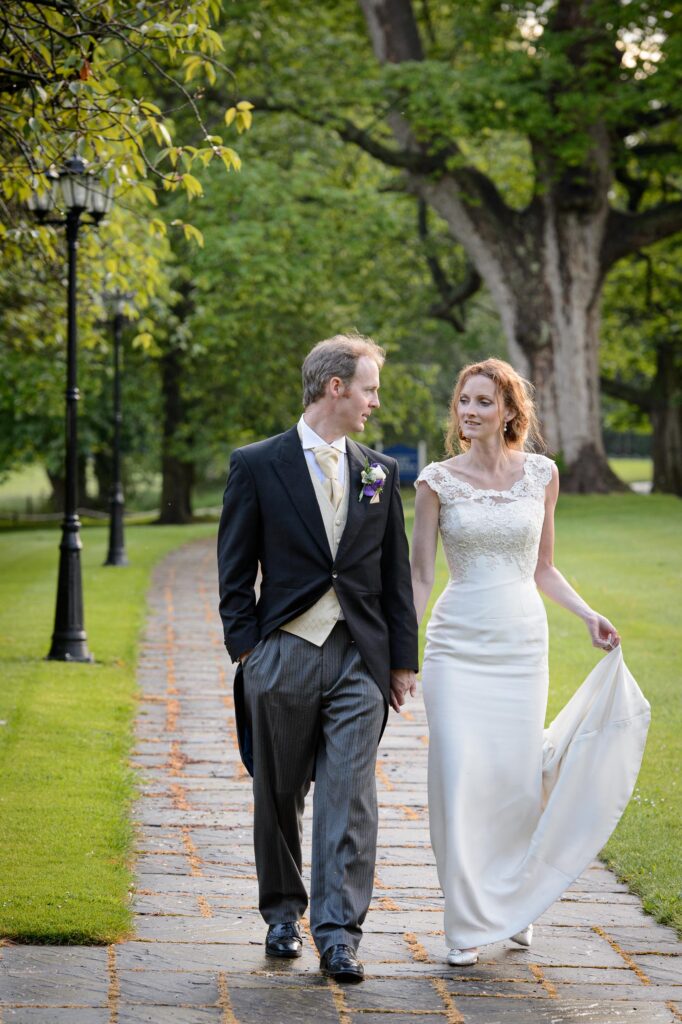 A bride and groom walking hand-in-hand down a stone path lined with green trees and vintage lamp posts at a Leeds wedding venue.