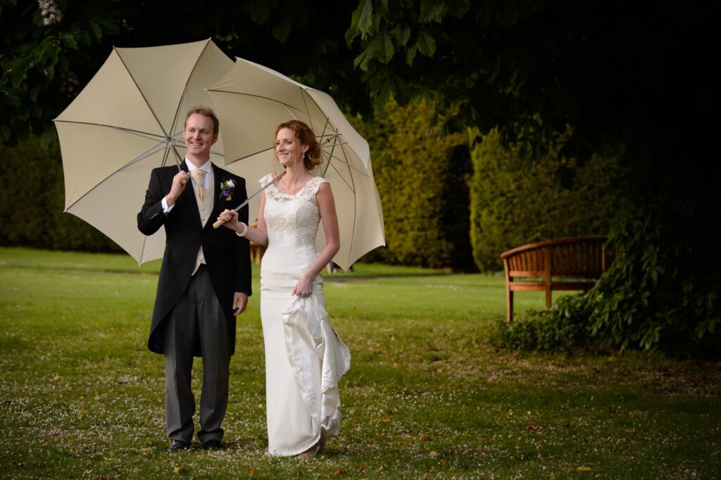 A bride in a lace wedding dress and a groom in a black morning suit standing on a green lawn under a large cream umbrella in Leeds.