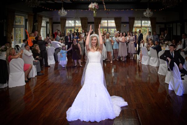A candid, full-view photo of a bride throwing a bouquet over her head into a crowd of people at a reception, all looking up, taken by a Leeds wedding photographer.