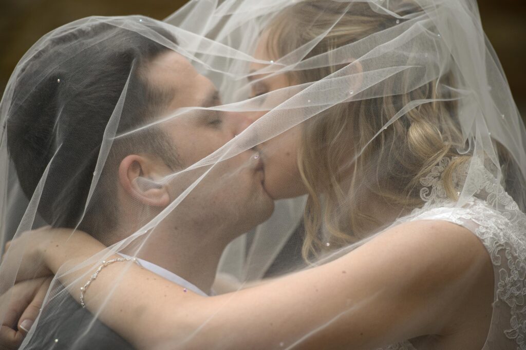 A very close, intimate portrait of a bride and groom kissing, partially covered and framed by a delicate, transparent wedding veil with bead details. Photo by a Leeds wedding photographer.