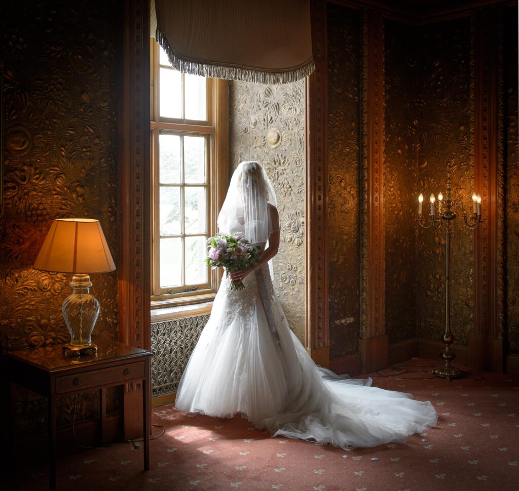 A full-length portrait of a bride in a long, dramatic veiled dress with an extensive train, standing near a window in a historic, richly wallpapered room, shot by a Leeds wedding photographer.