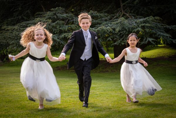 Children Playing: "A group of children in wedding attire—two girls in white dresses and a boy in a suit—holding hands and running across a green lawn."