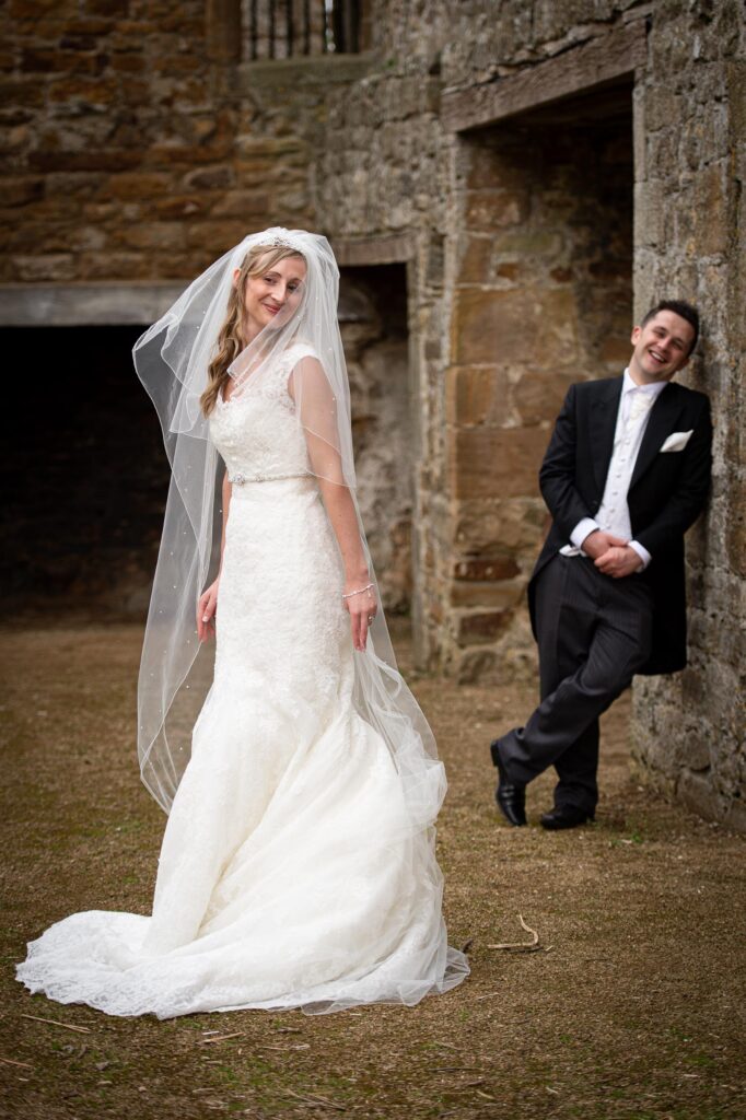 An outdoor portrait of a bride posing playfully for the camera while the groom leans casually against an ancient stone wall in the background, by a Leeds wedding photographer.