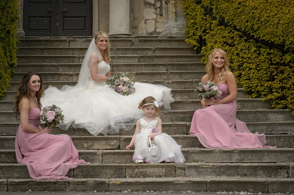 Group portrait of a bride, two bridesmaids in pink, and a flower girl in white, sitting on old stone steps outside a wedding venue, captured by a Leeds wedding photographer.