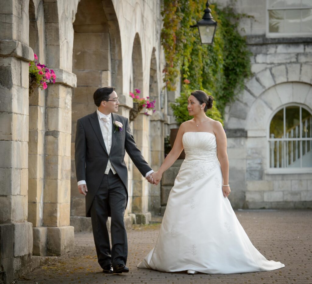 Bride and groom holding hands walking beside elegant stone arches at a Leeds wedding venue, photographed by Howard Barnett