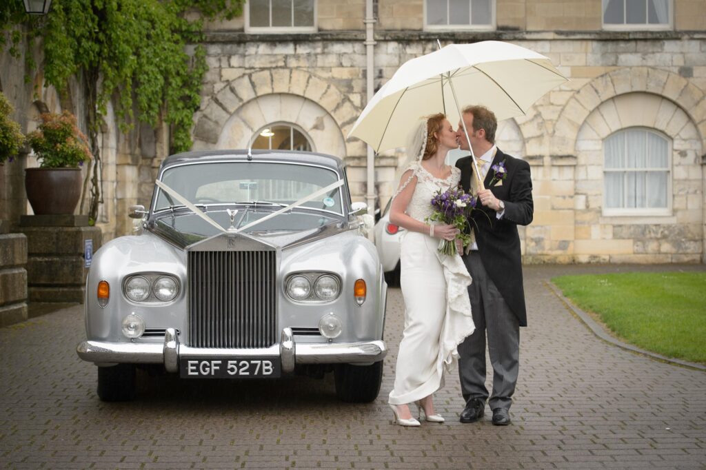 A bride and groom kissing under a white umbrella next to a silver vintage Rolls Royce with white ribbons in front of a grand stone building.