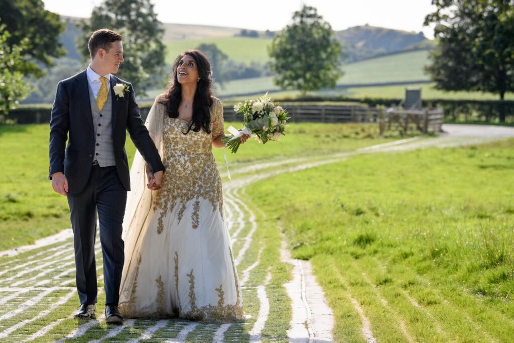 A bride in a gold-embroidered gown and a groom walking hand-in-hand through a green field on a sunny day in the Yorkshire countryside.