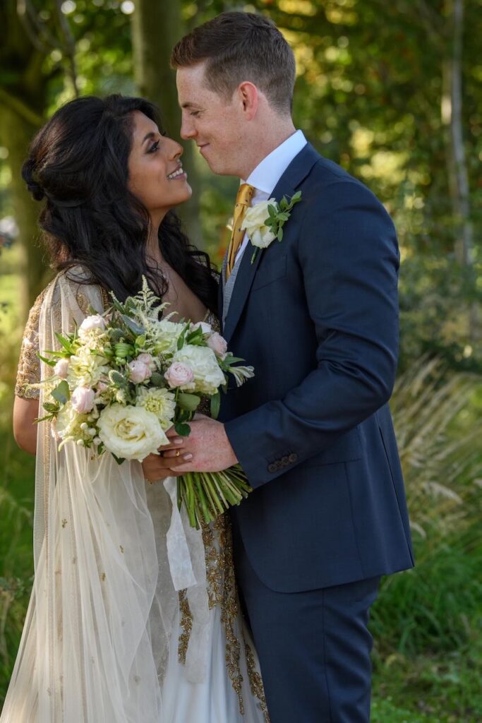 A close-up portrait of a bride holding a lush white and blush pink bouquet, standing next to the groom in a navy suit during a wedding in Harrogate.