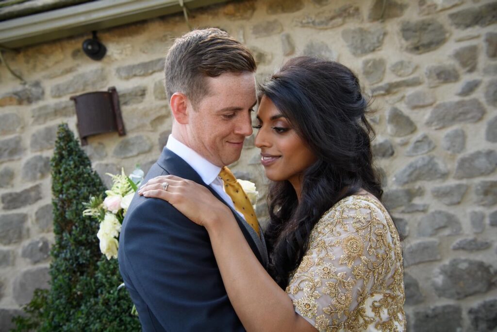 An intimate moment between a bride and groom leaning against a rustic stone wall, showcasing the bride's intricate gold beaded dress and engagement ring.